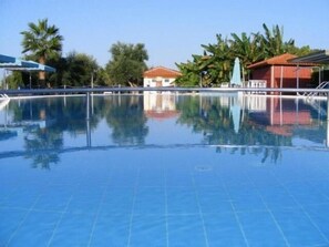 A clear blue swimming pool with a tiled deck, surrounded by palm trees and buildings with red roofs. - Lintzi (Pineios)