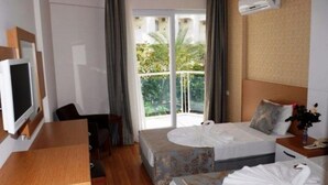 A hotel room with a bed, a desk, a chair, a TV, and a balcony with a view of greenery. - Blue Fish (Alanya)