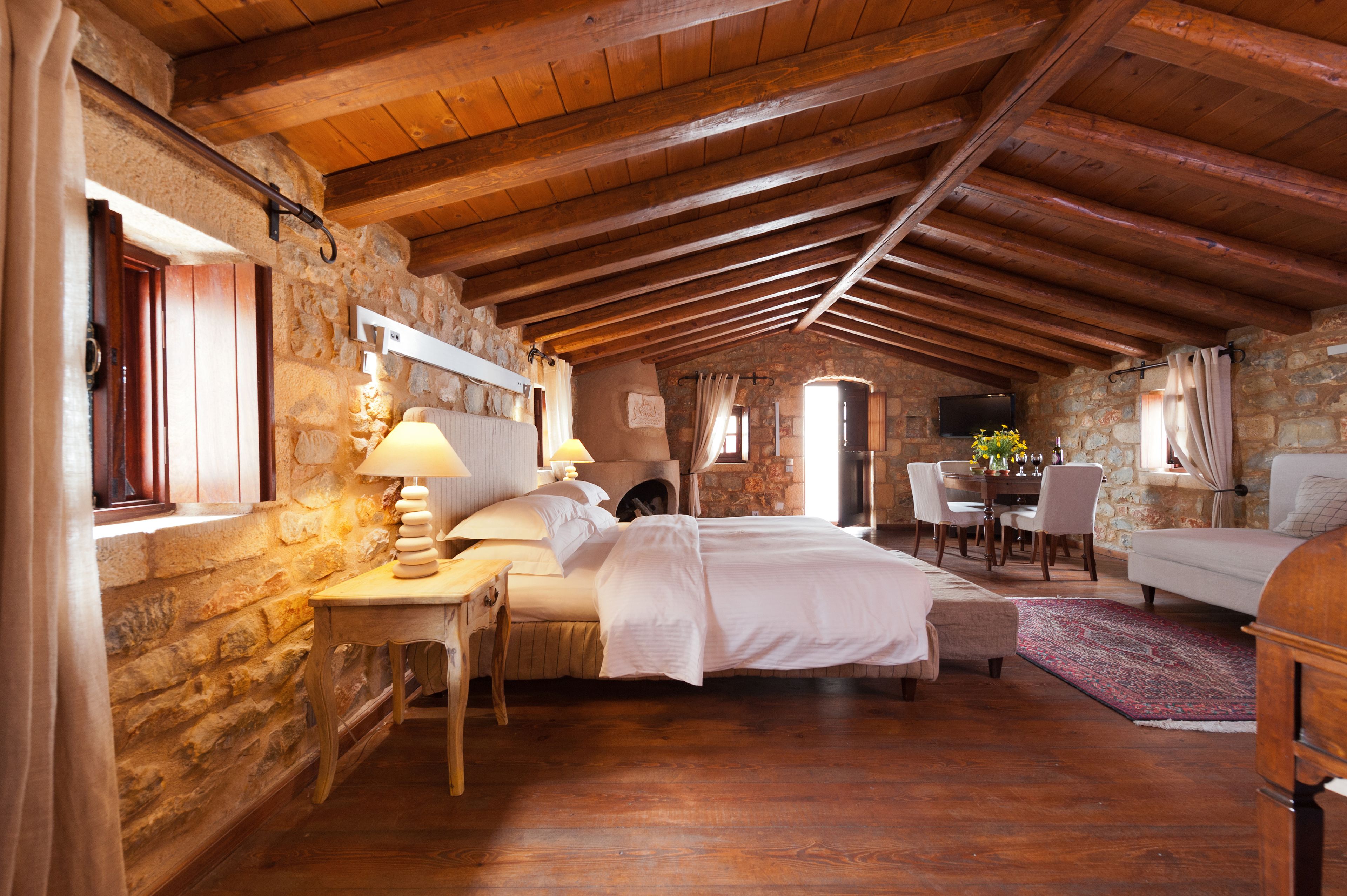 A rustic bedroom with a stone wall, wooden ceiling beams, a bed, a desk with a lamp, a chair, and a TV on the wall.