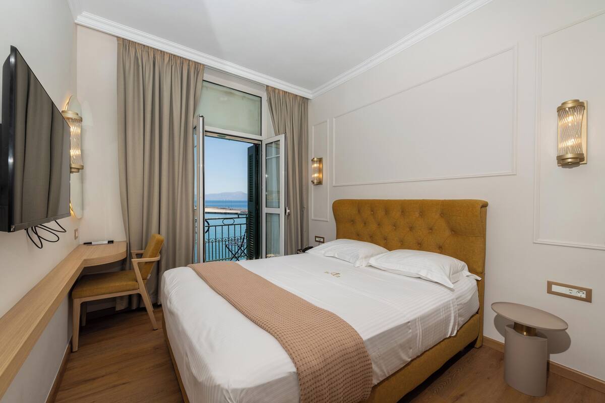 Une chambre d’hôtel comprenant un lit, un bureau, une chaise, une télévision, une fenêtre avec des rideaux et un balcon.