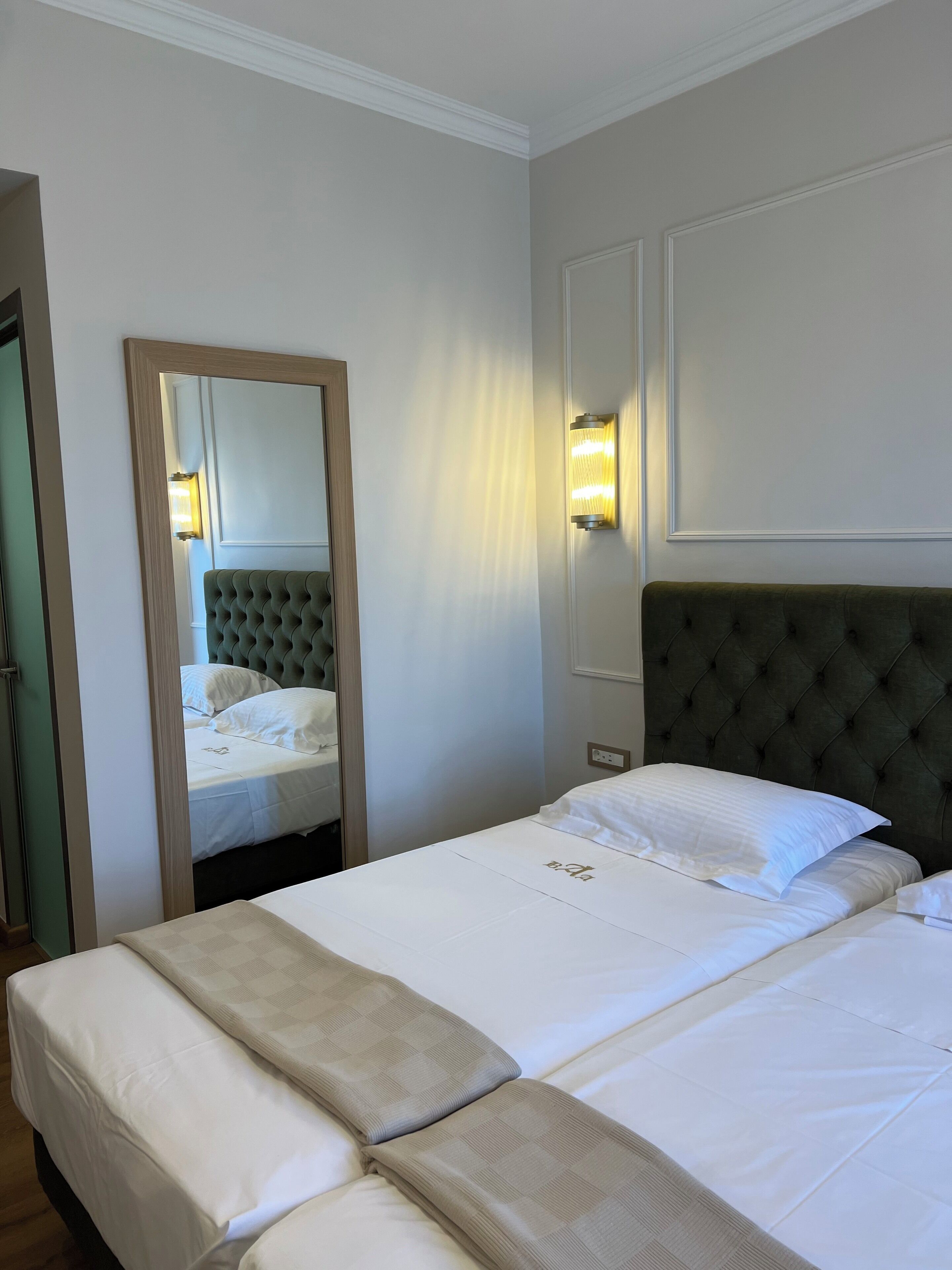 A hotel room with a large bed, a mirror, a wall-mounted light, and a door.