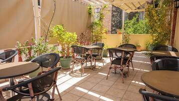 An outdoor patio area with several tables and chairs, surrounded by potted plants and a pergola structure.