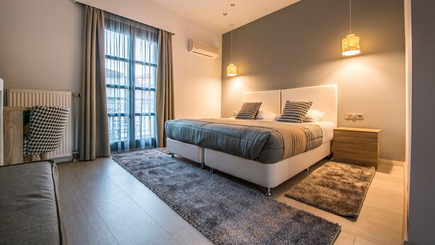 Une chambre moderne avec un grand lit, une table de chevet en bois, un radiateur, un fauteuil à motif pied-de-poule et une vue sur l’extérieur par une fenêtre.