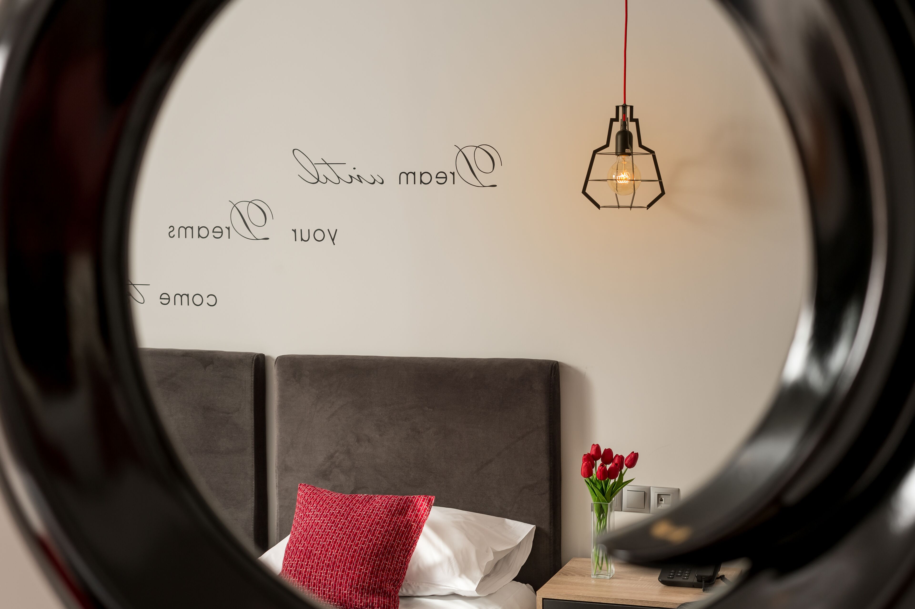 A bedroom with a bed, a nightstand, a vase with flowers, and a wall with a light fixture and text.