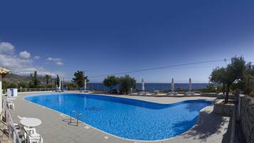 A swimming pool with a curved edge, surrounded by lounge chairs and umbrellas, with a view of the sea and mountains in the background.
