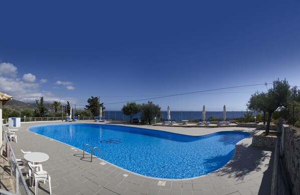 A swimming pool with a curved edge, surrounded by lounge chairs and umbrellas, with a view of the sea and mountains in the background. - Kardamili Beach Hotel (West Mani)