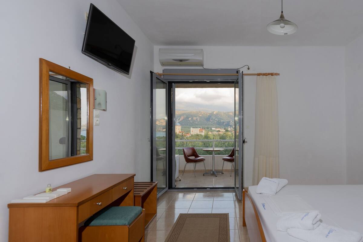 A hotel room with a bed, a desk, a chair, a TV, and a balcony with a view of the mountains.