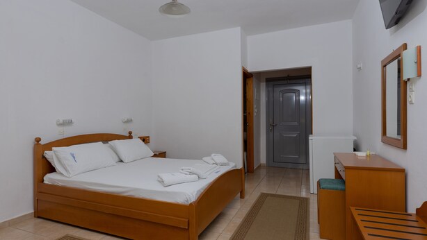 A neatly made bed with white linens and pillows, a wooden bedside table with a lamp, and a small desk with a mirror and a bottle. - Kardamili Beach Hotel (West Mani)