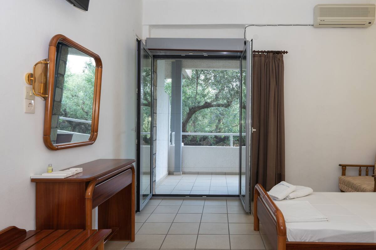 A hotel room with a wooden bed, a wooden nightstand, a mirror, a sliding glass door leading to a balcony, and an air conditioning unit.