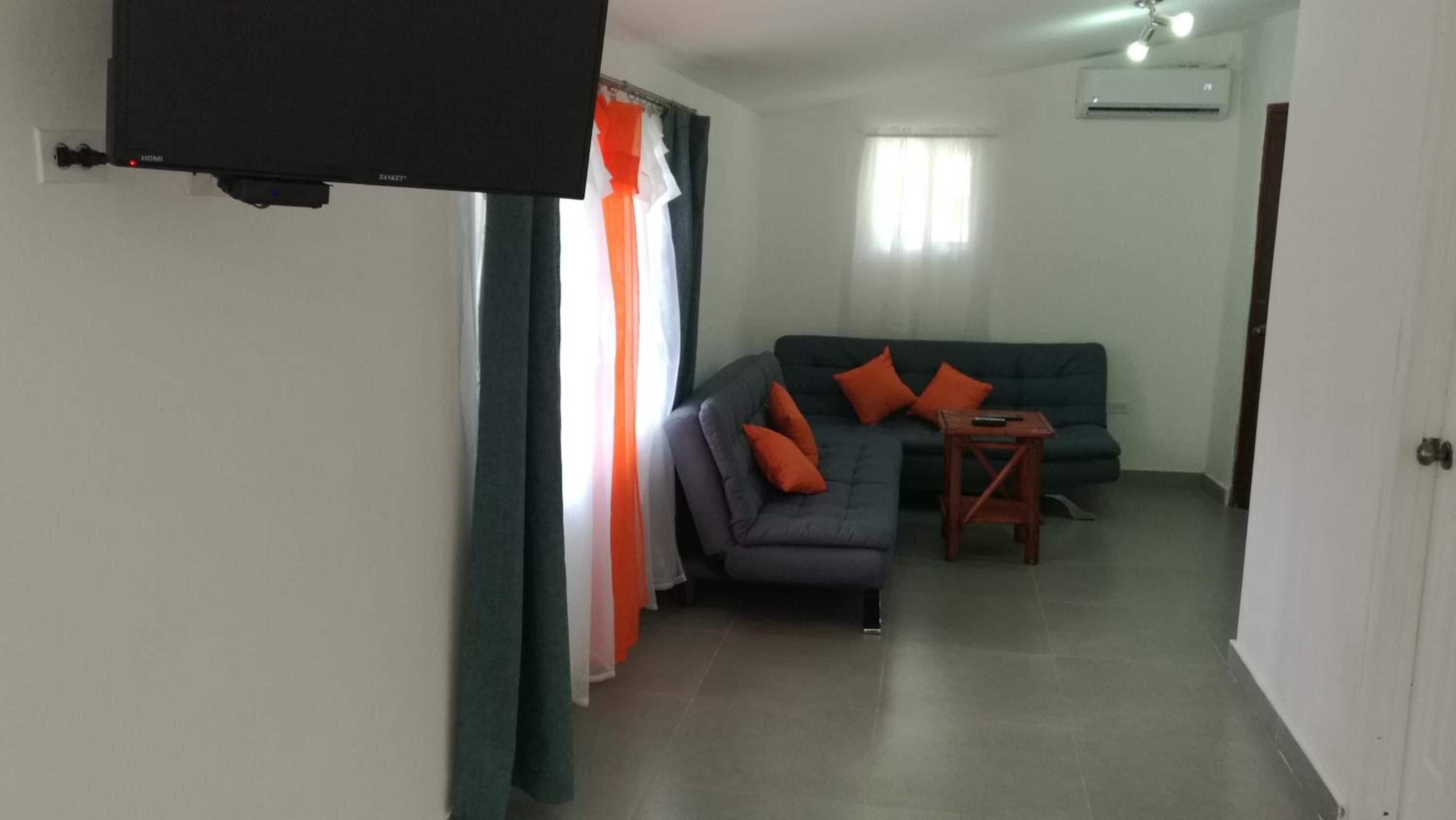 A living room with a grey sofa, orange cushions, a small wooden table, and a flat-screen TV mounted on the wall.