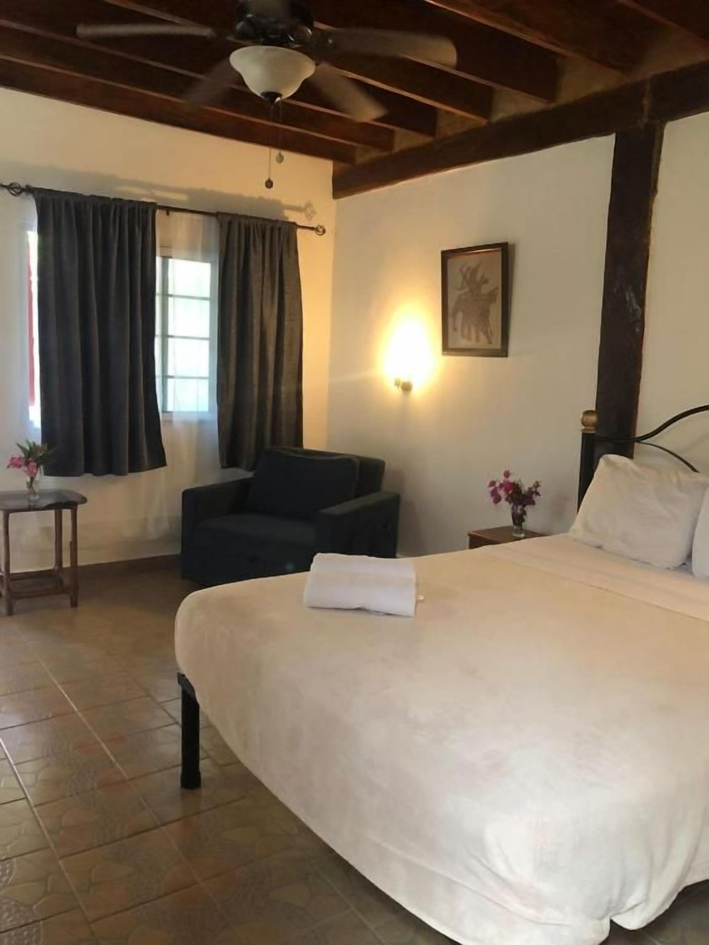 A bedroom with a large bed, a chair, a small table, and a wall-mounted light.