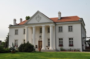 Un edificio grande y blanco con una fachada simétrica, destacando una entrada central flanqueada por columnas, un frontón triangular y un escudo sobre la entrada.