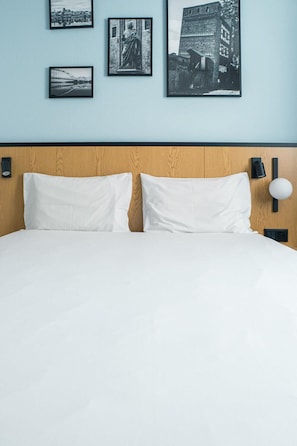 Una cama con ropa de cama blanca, cabecera de madera e iluminación montada en la pared en color negro.