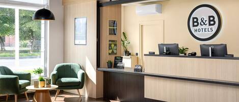 A reception area with a B&B Hotels sign, a reception desk, two green chairs, a small table, and a potted plant.