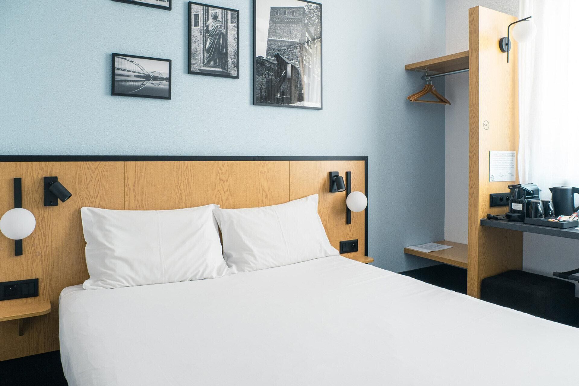 Habitación de hotel con cabecera de madera, ropa de cama blanca y tres fotografías blanco y negro en la pared.
