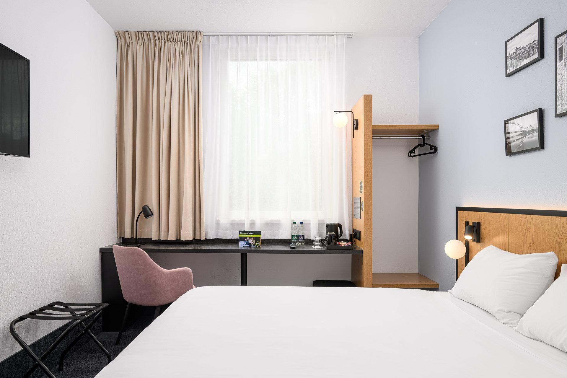 Een hotelkamer met een bed, een bureau met stoel, een raam met gordijnen, een wandlamp en ingelijste foto's.