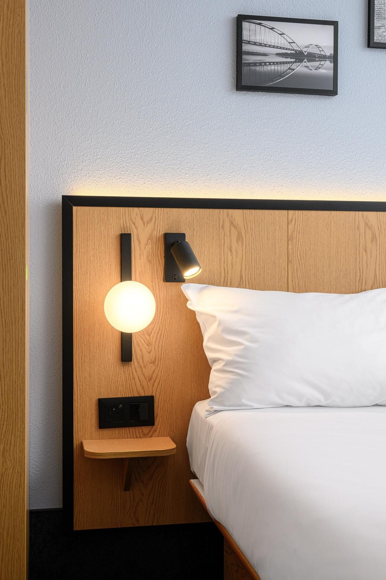 A hotel room with a wooden headboard, a bedside table with a lamp, and a bed with white bedding.