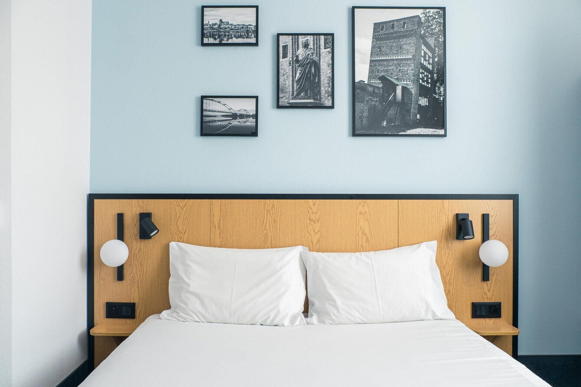 Ein Hotelzimmer mit einem Holz-Kopfteil, weißer Bettwäsche und drei Schwarz-Weiß-Fotos an der Wand.