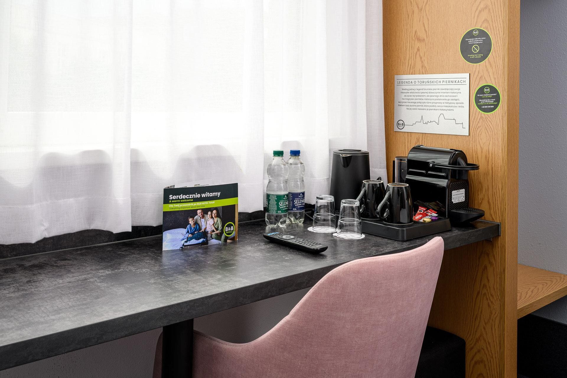 Ein Hotelrezeptionstresen mit einer Kaffeemaschine, Wasserflaschen, einer Fernbedienung und einer Willkommensbroschüre.