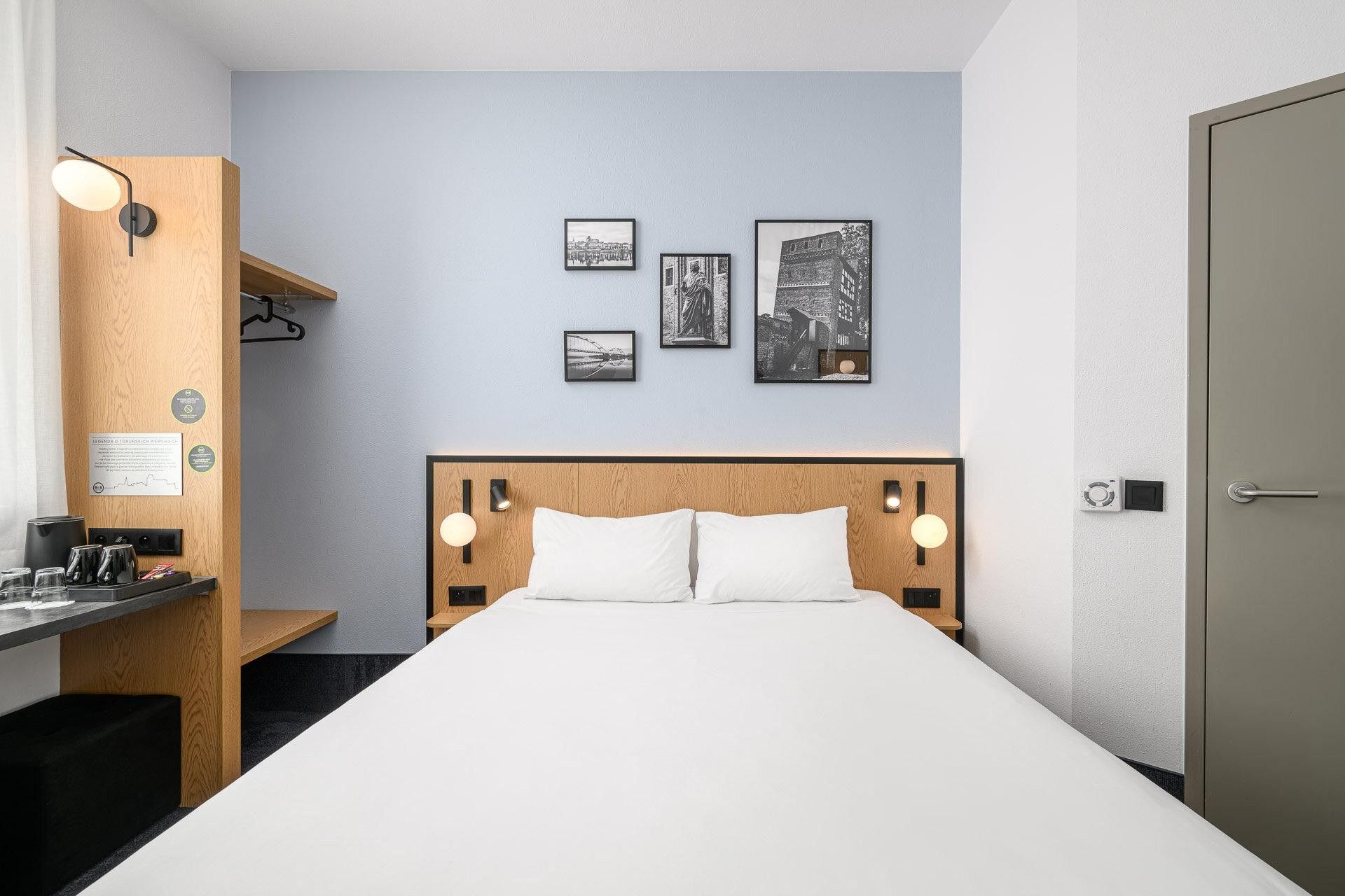 Ett hotellrum med en säng, sänglampor, en sänggavel i trä, ett skrivbord med en lampa och konstverk som hänger på väggen.