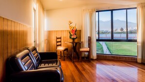 A room with a wooden floor, a leather sofa, a small table with chairs, and a large window with a view of mountains and a field. - DM Hoteles Arequipa (Arequipa)
