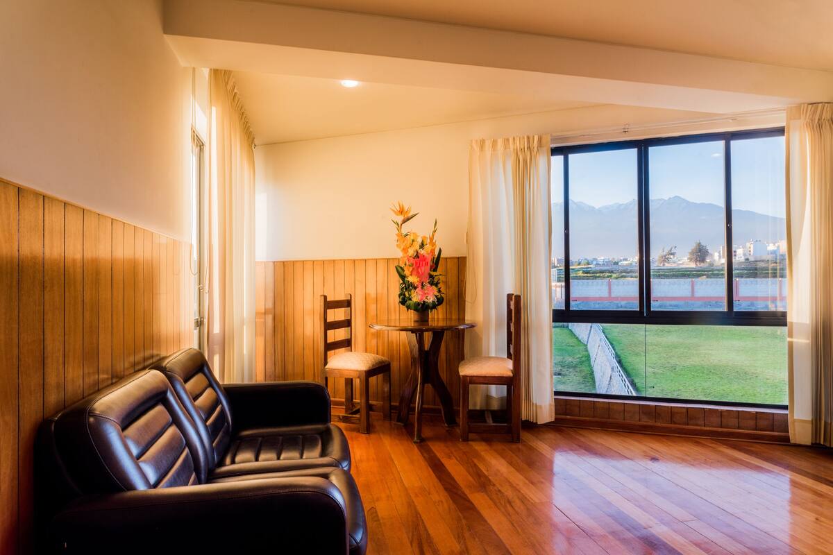 A room with a wooden floor, a leather sofa, a small table with chairs, and a large window with a view of mountains and a field.