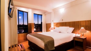 A hotel room with a large bed, wooden headboard, a bedside table with a lamp, a nightstand with a phone, and a window with curtains. - DM Hoteles Arequipa (Arequipa)