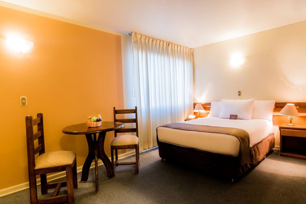 A hotel room with a bed, a desk with a chair, a small table with fruit, and a window with curtains.