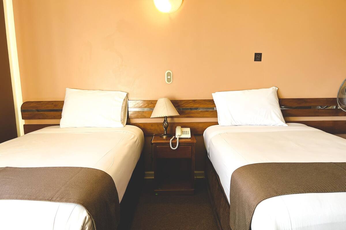 A hotel room with two single beds, a bedside table with a lamp and phone, and a wall-mounted light fixture.