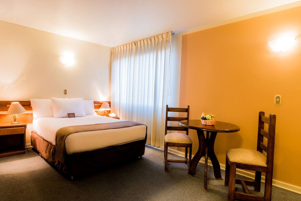 A hotel room with a bed, bedside tables, a desk with a chair, and a window with curtains.