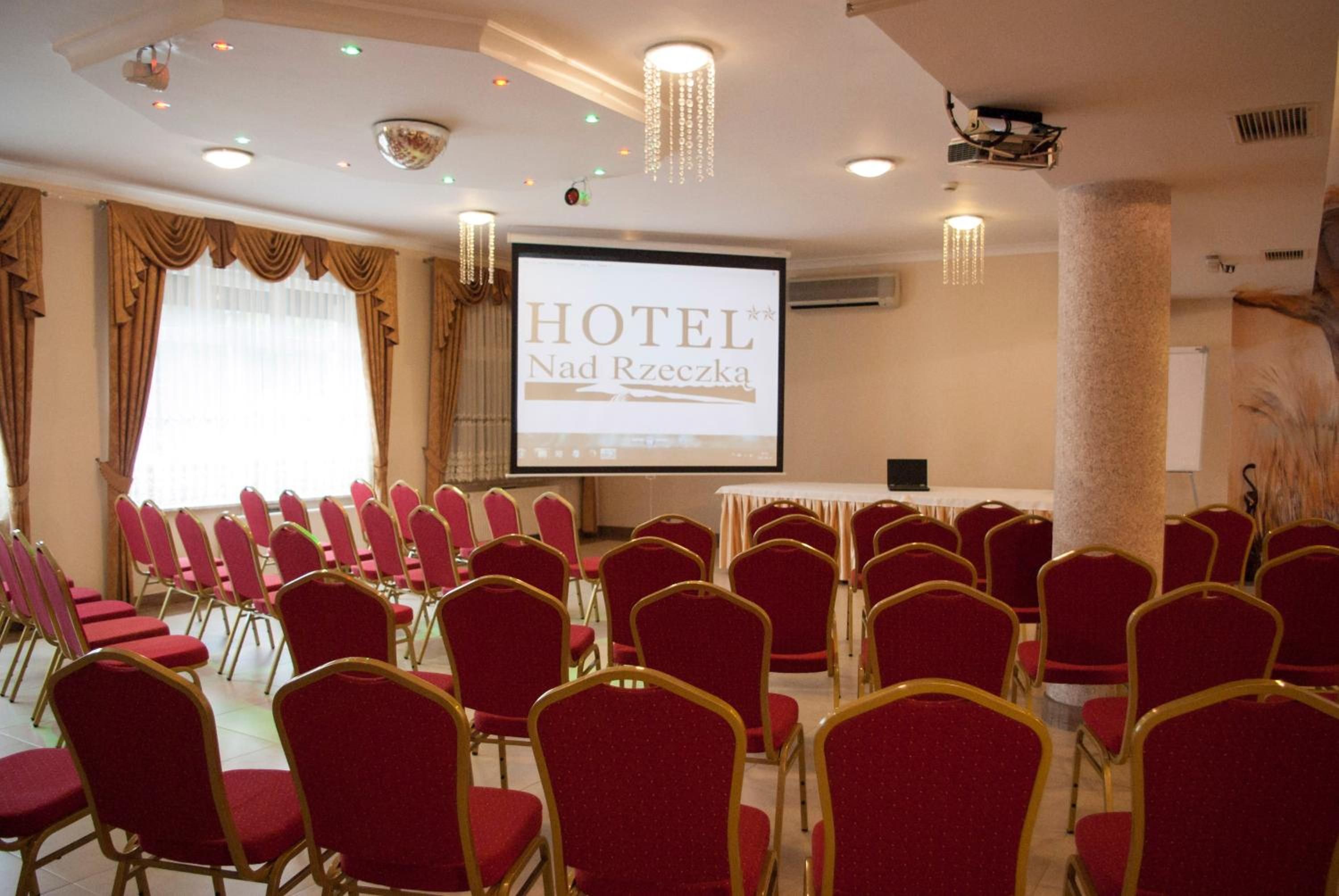A hotel conference room with rows of red chairs, a projector screen displaying 'HOTEL Nad Rzeczką', and a large pillar.