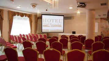 A hotel conference room with rows of red chairs, a projector screen displaying 'HOTEL Nad Rzeczką', and a large pillar.