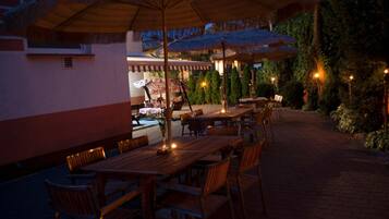 An outdoor dining area with wooden tables and chairs, umbrellas, and a pool in the background.