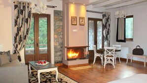 A cozy room with a fireplace, wooden flooring, a dining area with white chairs, and a large window with curtains.