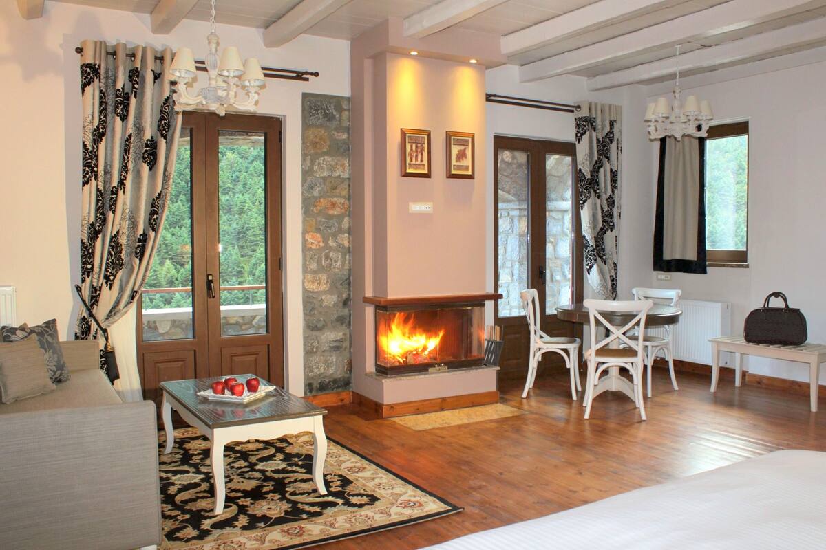Une pièce chaleureuse avec une cheminée, un plancher en bois, un coin repas avec des chaises blanches et une grande fenêtre avec des rideaux.