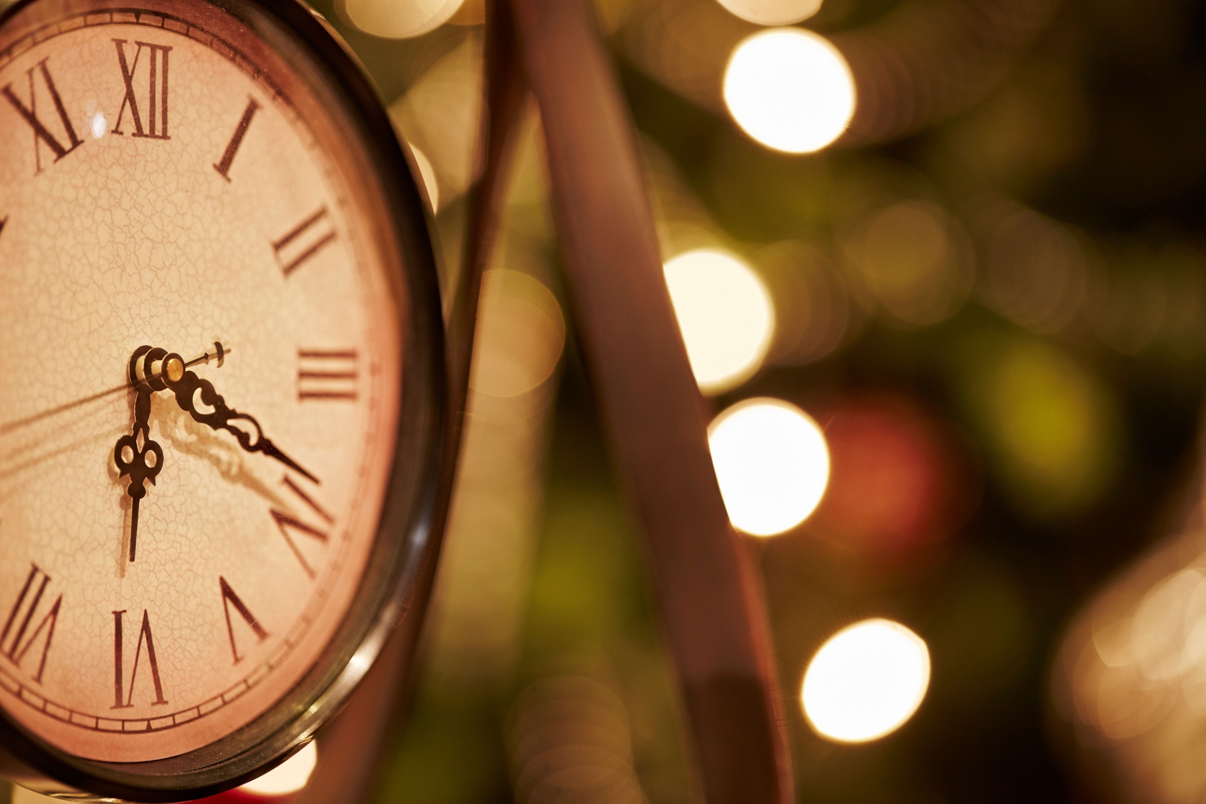 A close-up of a clock with Roman numerals, set against a blurred background with bokeh lights.