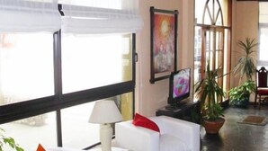 Un lobby amplio con sofás blancos, una mesa con lámpara, ventanas grandes y plantas en macetas.