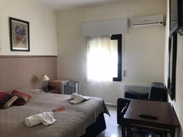 A hotel room with a bed, a desk, a chair, a lamp, an air conditioning unit, a window with curtains, and a framed picture on the wall.