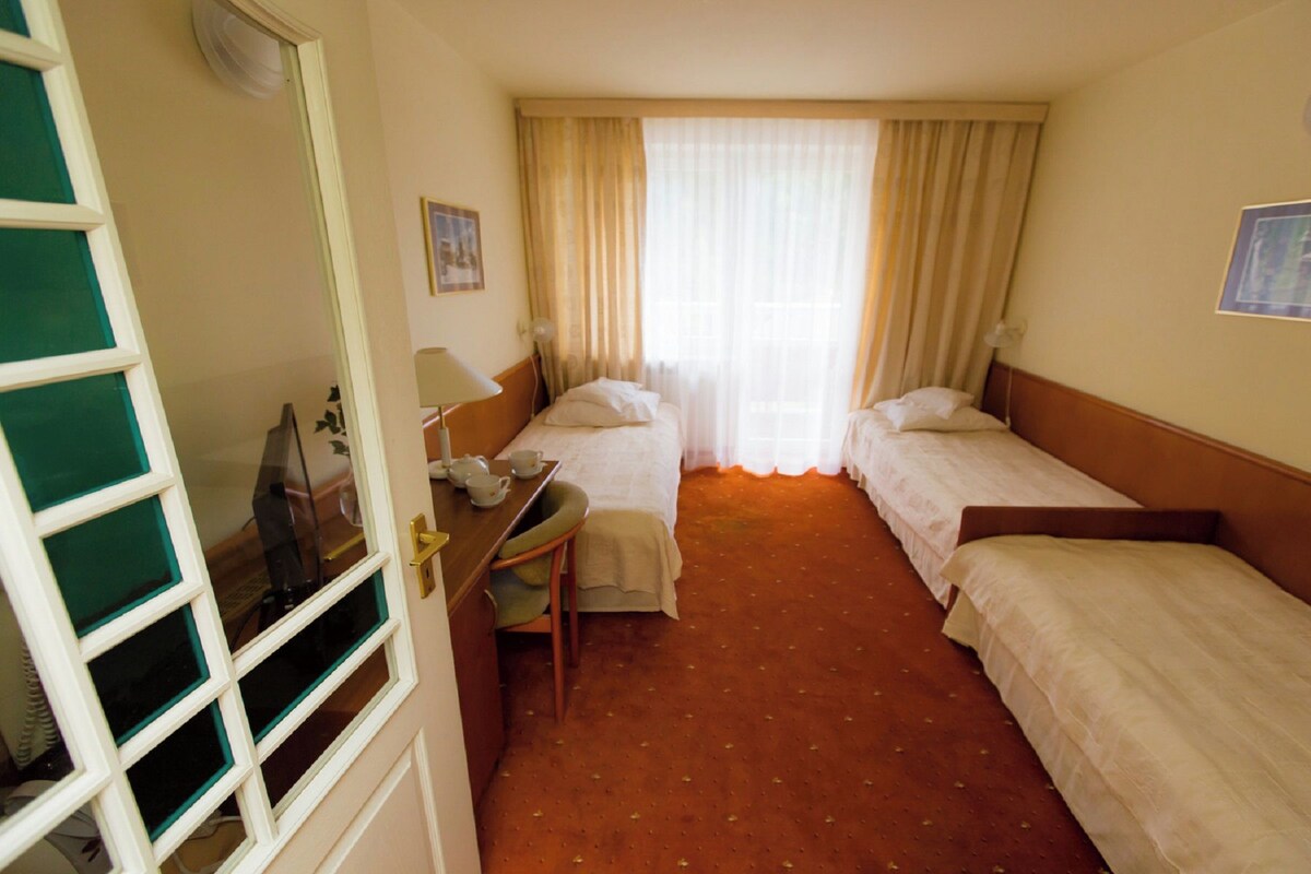 A hotel room with two single beds, a desk with a chair, a small table with cups and a lamp, and a window with curtains.