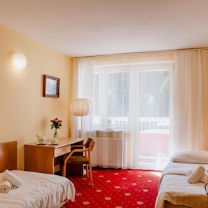A hotel room with a bed, a desk with a chair, a lamp, a window with curtains, and a view of the outside. - Geovita Krynica Zdroj (Krynica-Zdroj)