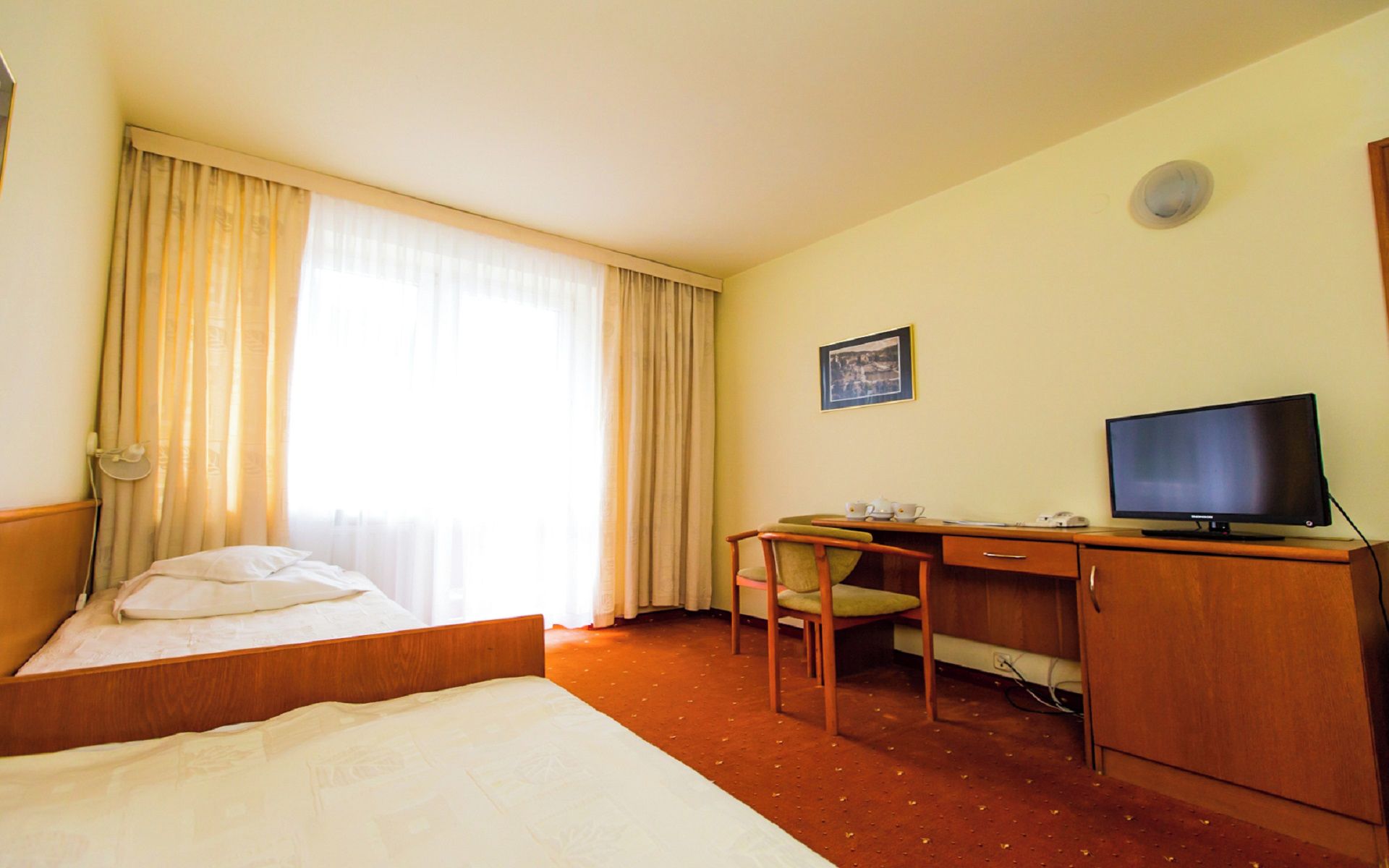 A hotel room with a bed, a desk with a chair, a television, and a window with curtains.