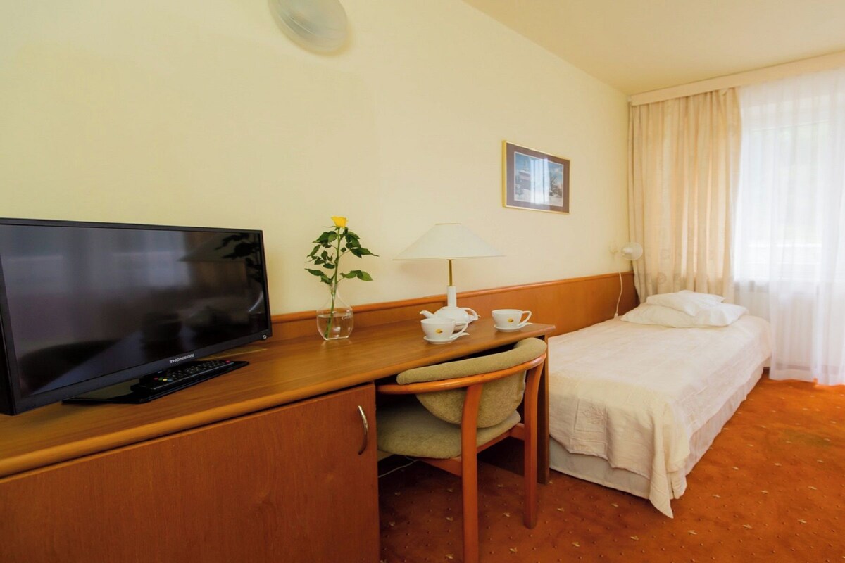 A hotel room with a television, a desk with a lamp, a chair, a bed, and a window with curtains.