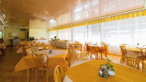 A dining area with multiple tables and chairs, large windows with sheer curtains, and a kitchen area in the background. - Geovita Krynica Zdroj (Krynica-Zdroj)
