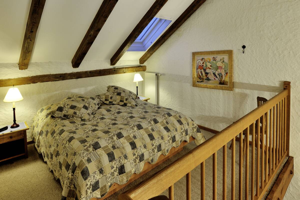 Ein Schlafzimmer mit schrägem Dach, Holzbalken, einem Bett mit kariertem Überwurf, zwei Nachttischlampen, einem Bild an der Wand und einem hölzernen Geländer einer Treppe.