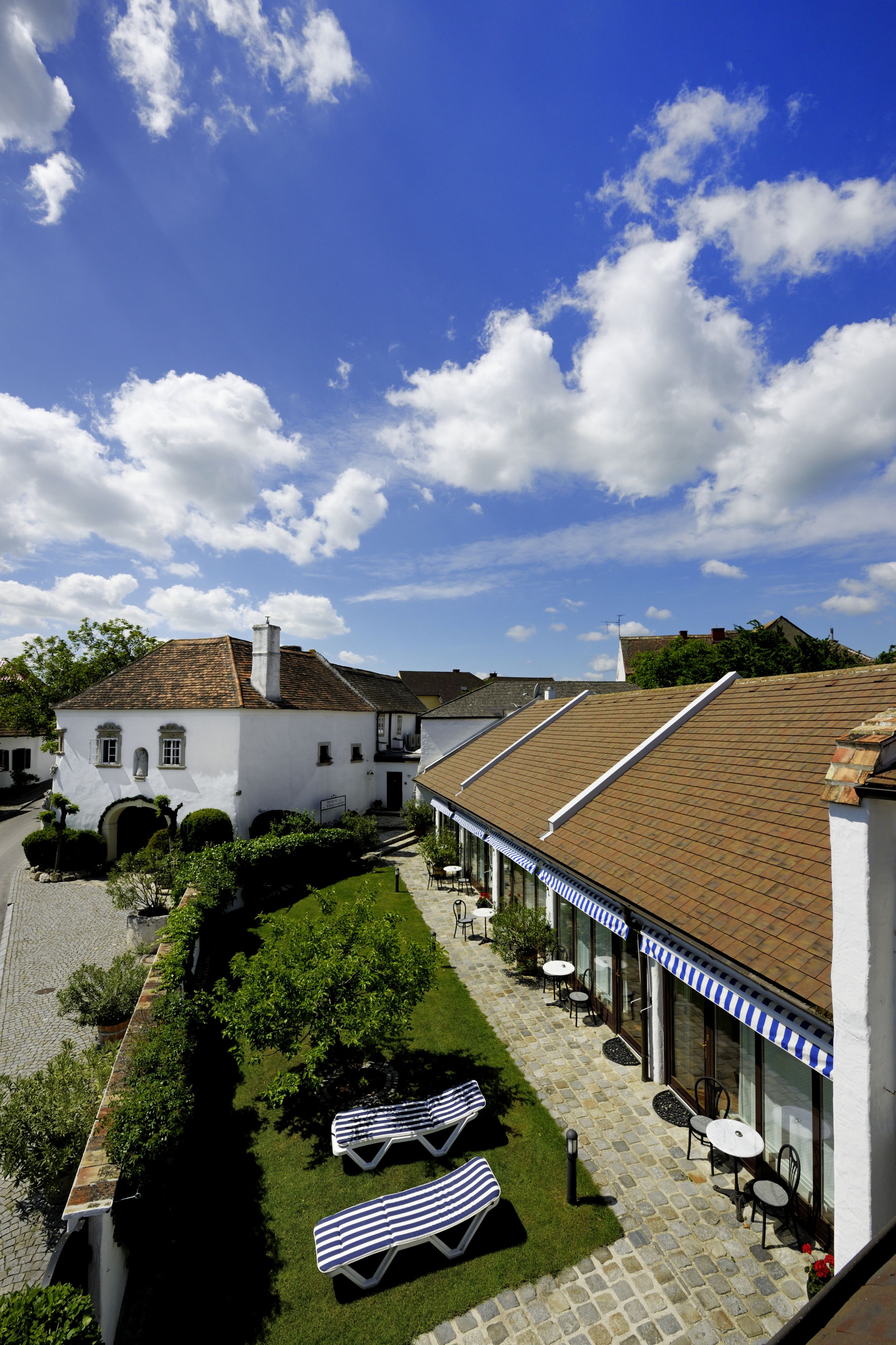 A courtyard with lounge chairs and tables, a white building with a tiled roof, and a clear blue sky with clouds.
