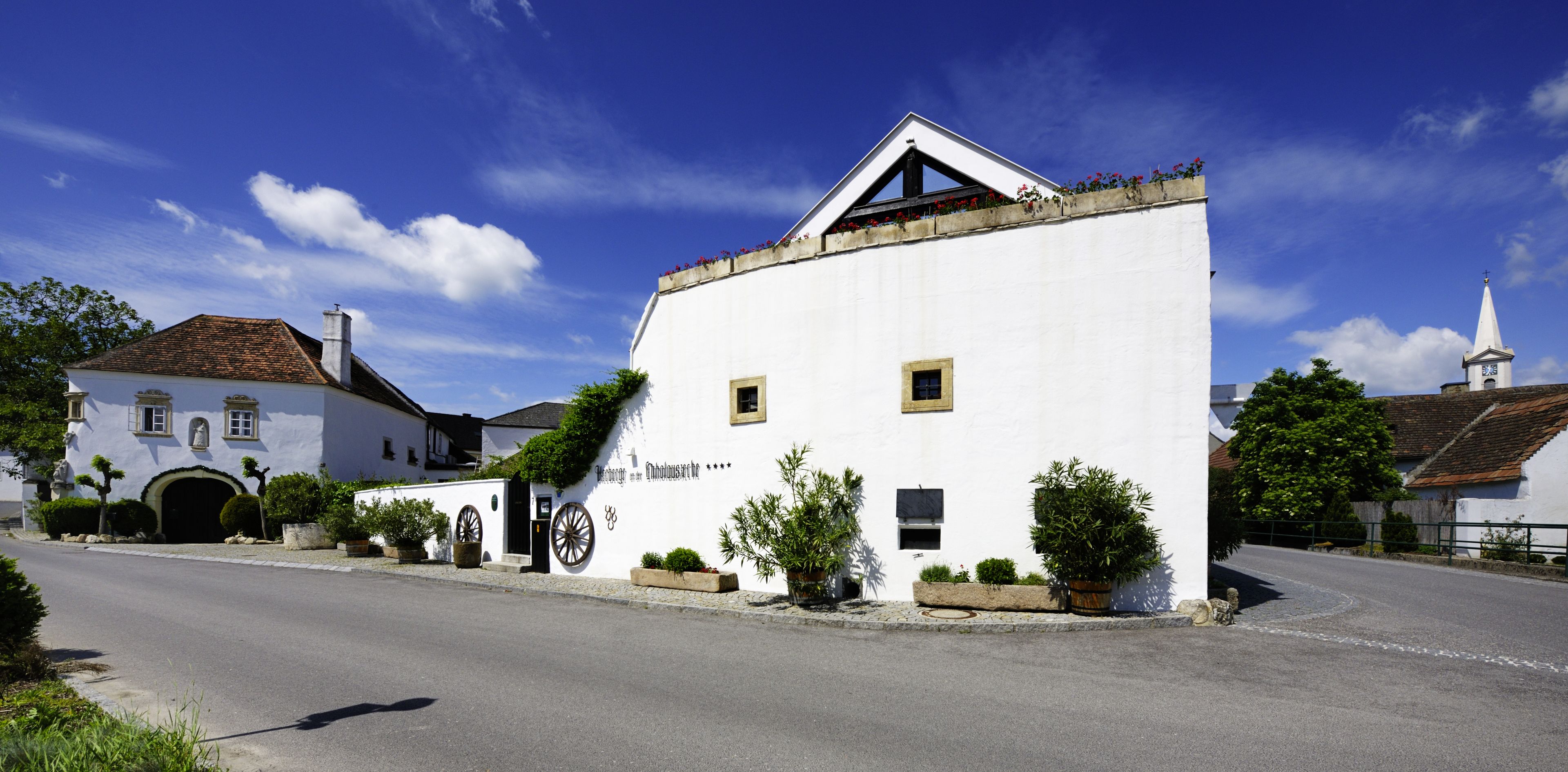 A white building with a traditional design, featuring a small balcony with flowers, a clock tower, and a road in front.