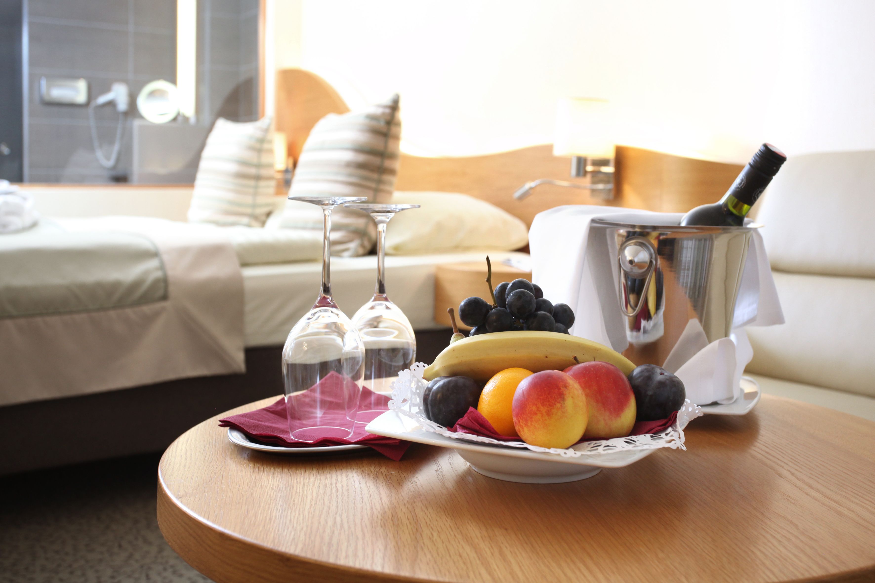 Una habitación de hotel con cama, una mesita de noche con frutero, dos copas de vino y una botella de champán.