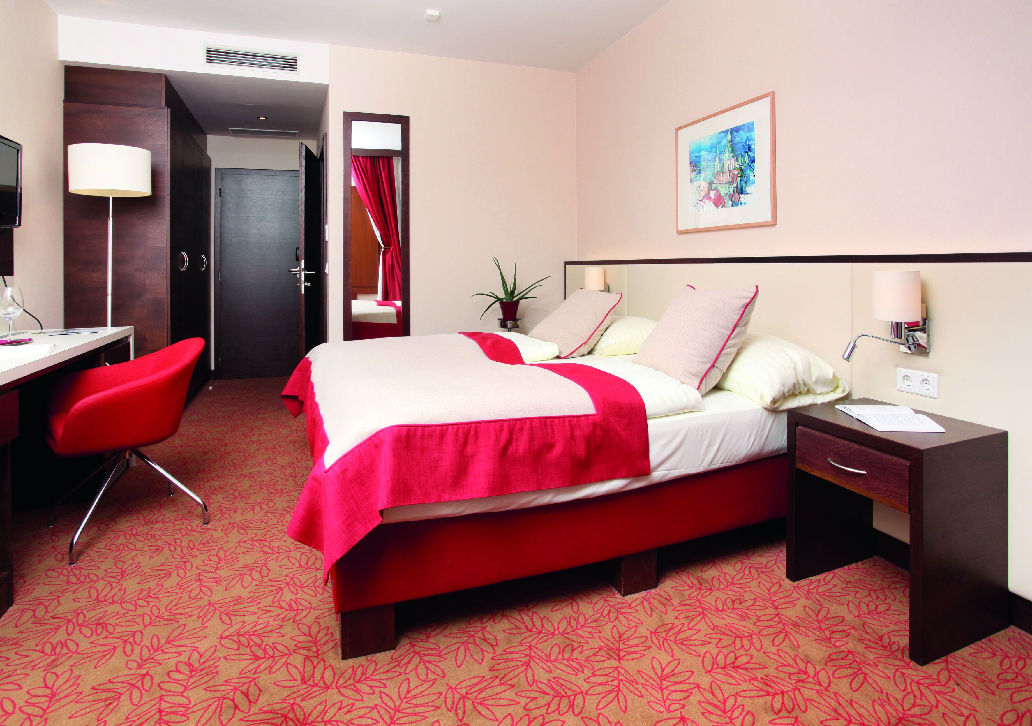 A hotel room with a bed, a desk with a chair, a nightstand, a lamp, and a painting on the wall.