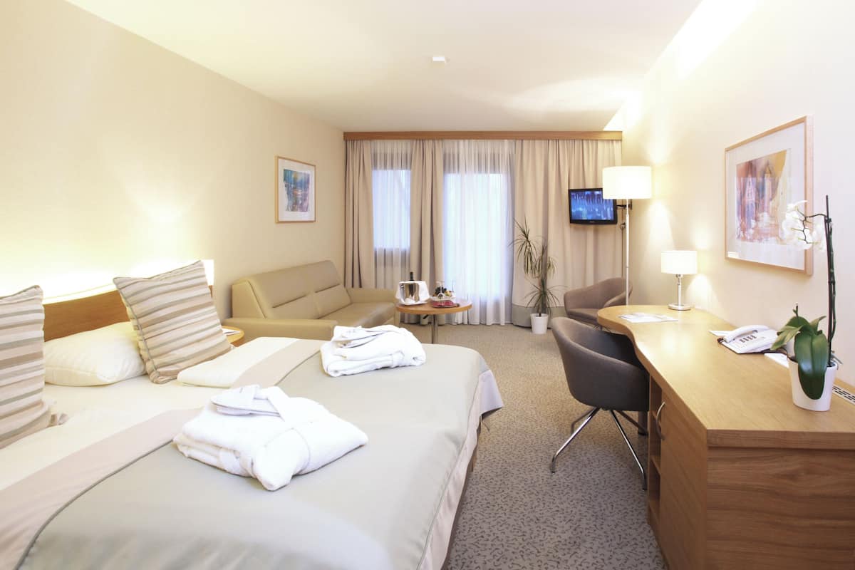 Una habitación de hotel con cama, escritorio, silla, televisor y ventana con cortinas.