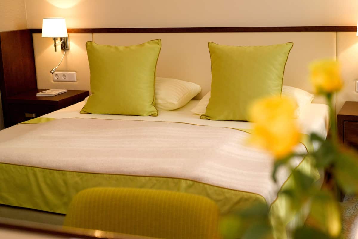 Habitación de hotel con una cama, dos almohadas verdes, una almohada blanca, una mesita de noche con una lámpara y una silla amarilla.